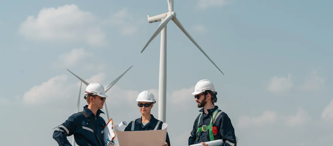 Engineering team at a wind farm Three engineers in protective gear reviewing plans beneath wind turbines at a wind farm